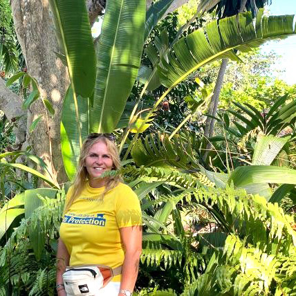 Blonde woman standing in front of tropical green plants wearing a yellow Cats Protection t-shirt
