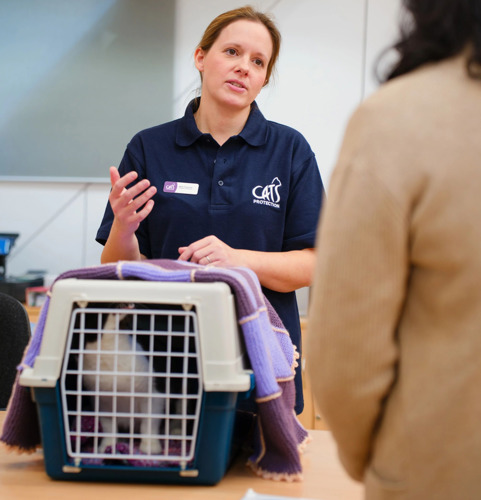 A woman with brunette hair tied back and wearing a navy blue t-shirt featuring the Cats Protection logo stands behind a cat carrier resting on a table. The carrier is made from blue and white hard plastic, opens from the front and top, has a black-and-white cat inside partially covered with a stripy purple blanket