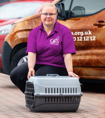 A woman with blonde hair tied back, glasses and a pink t-shirt featuring the Cats Protection logo is crouched down on paving outdoors with her hands resting on an empty cat carrier. The carrier is made from hard grey plastic, opens from the front and top and has vents on the side.