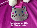 Image of a cat shaped medal lying on pink fabric with white cat paws visible above the medal. The text "I'm taking on 50k in May for cats" overlays the image.