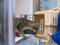 A brown tabby cat peering into an open, beige, plastic cat carrier being held by a person wearing a blue jumper